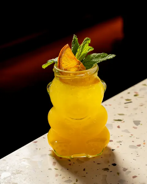 Golden slushy drink in a bear-shaped glass with a dried orange slice and mint, on terrazzo
