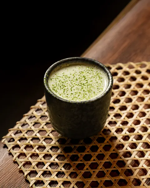Frothy matcha drink dusted with green powder in a dark speckled stoneware cup on a bamboo mat