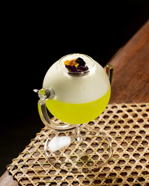 Neon green drink under white foam in a tilted globe glass, topped with pansies, on a bamboo mat