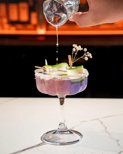 Clear liquid poured into a lavender cocktail with a sugared rim and cucumber ribbon in a scalloped coupe