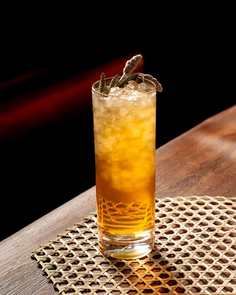 Tall amber highball over crushed ice with dried herb sprigs, lit by warm side light on a bamboo mat