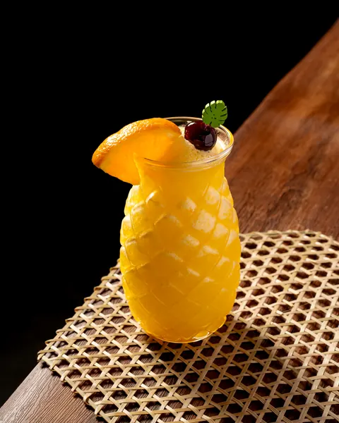 Frozen yellow drink in a pineapple-shaped glass with an orange wedge, cherry and monstera-leaf pick