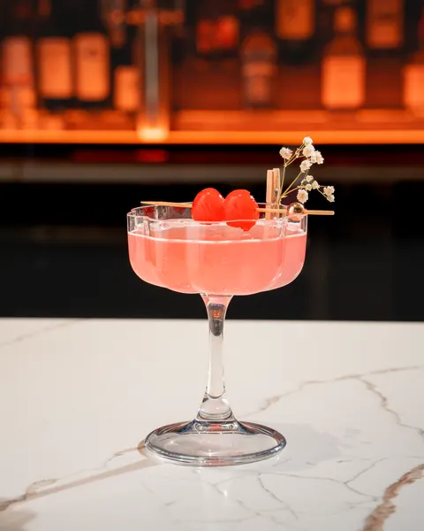 Pink cocktail in a scalloped coupe with two cherries and baby's breath, on marble before a glowing back bar