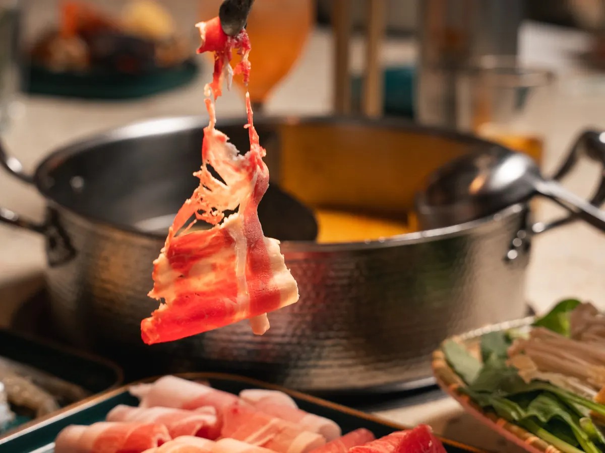 Tongs lift a thin slice of raw meat over a split hot pot of golden broth
