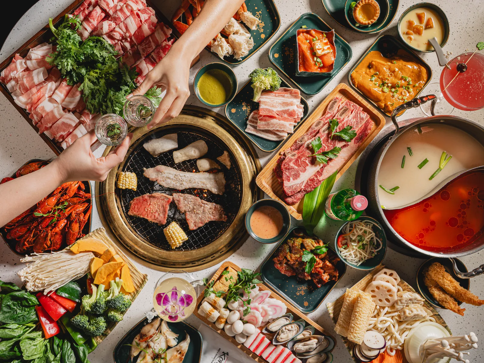 Overhead view of a full Kanpai table: a grill of short rib and corn, a split hot pot of white and red broth, platters of sliced pork and beef, crawfish and seafood