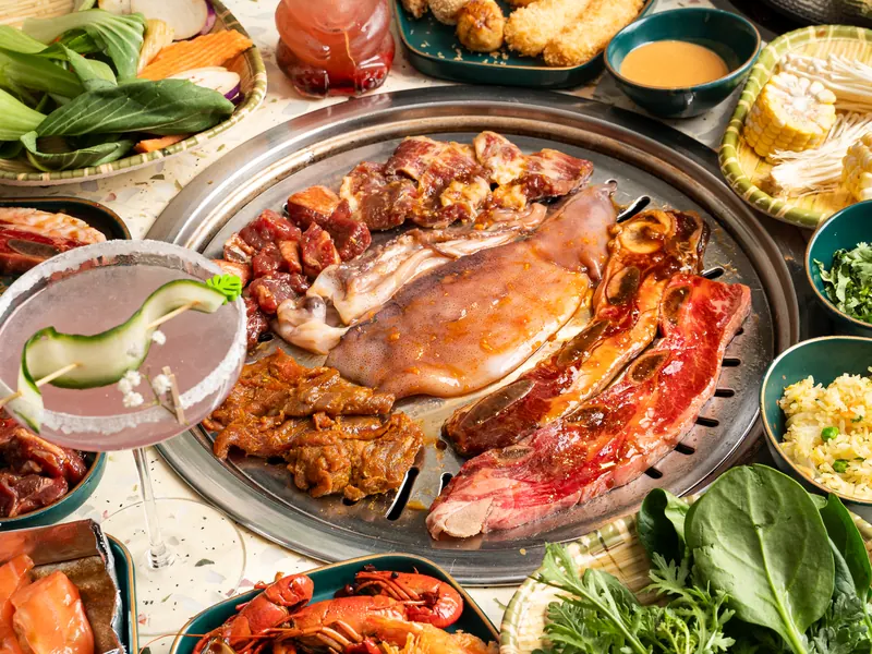 A round grill of marinated meats, squid and short ribs beside a cucumber-garnished cocktail, greens and corn