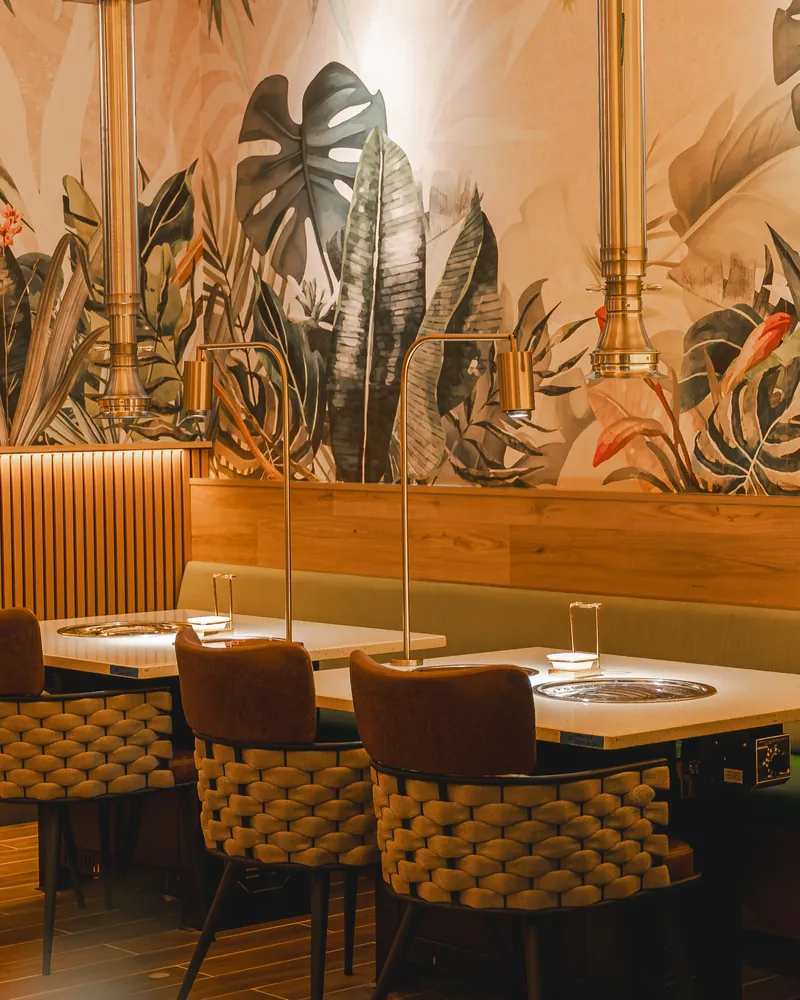 Grill tables with brass hoods in front of a tropical banana-leaf and monstera mural on a warm wall