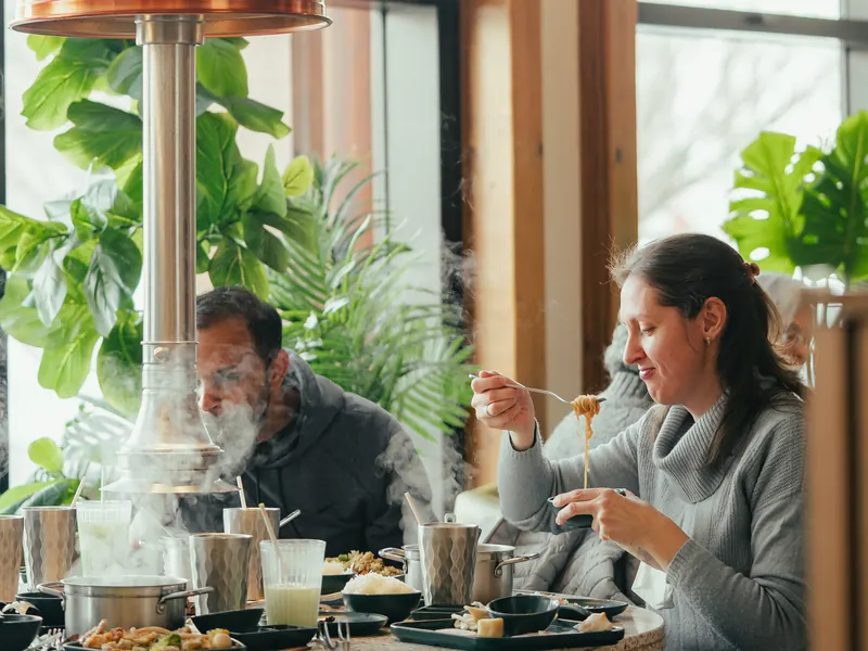 A guest twirls noodles at a sunny window table beside a steaming hotpot, framed by monstera and palms